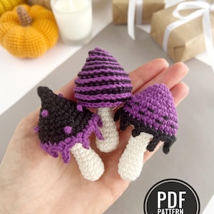 May include: Three small crocheted mushrooms with white stems and caps in purple and black. One cap is purple with black stripes, one is black with purple spots, and one is purple with black accents. A circular badge reads PDF PATTERN IN ENGLISH.