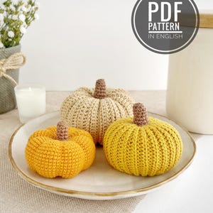 May include: Three crocheted pumpkins in shades of beige, yellow, and mustard, each featuring a brown stem. The pumpkins are arranged on a white plate. A circular graphic in the upper right corner contains the text PDF PATTERN IN ENGLISH.