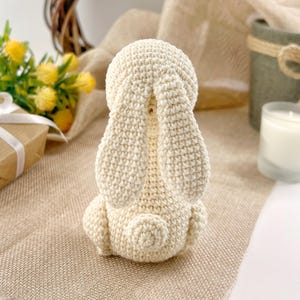 May include: A crocheted white bunny rabbit toy with long ears. The bunny is sitting on a beige textured surface.