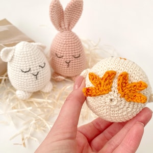 May include: Three crocheted stuffed animals: a pink bunny, a white lamb, and a white chick with orange wings. The chick is being held in a hand.