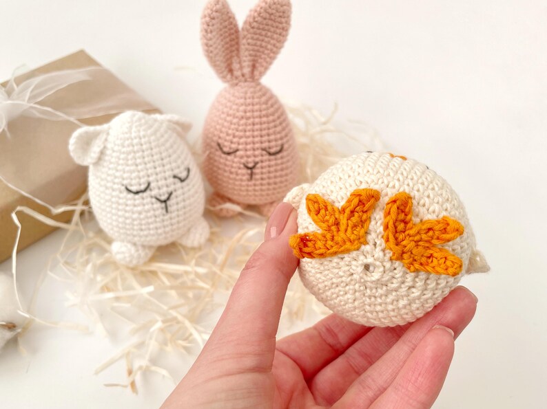 May include: Three crocheted stuffed animals: a pink bunny, a white lamb, and a white chick with orange wings. The chick is being held in a hand.