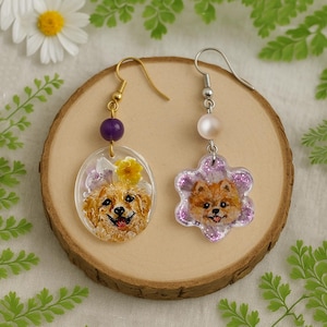 Pode incluir: Brincos feitos à mão com retratos de cães. Um brinco tem um pingente oval com um cão dourado, uma conta roxa e uma flor amarela. O outro tem um pingente em forma de flor com um retrato de cão Pomerânia e uma pérola.