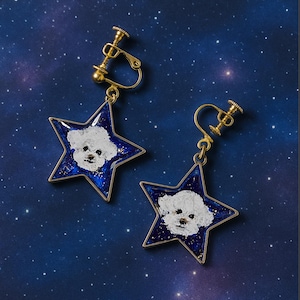 Pode incluir: Um par de brincos dourados de pressão. Cada brinco apresenta um pingente azul em forma de estrela com um desenho de rosto de cão branco. Os pingentes de estrela têm uma borda dourada e estão sobre um fundo azul escuro com glitter.