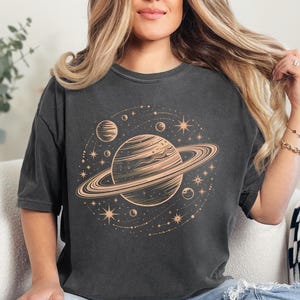 Könnte beinhalten: Dunkelgraues T-Shirt mit einem Himmelsdesign. Die Grafik zeigt einen Planeten mit Ringen, andere Planeten, Sterne und Sternbilder in Beige. Das T-Shirt hat kurze Ärmel und eine lockere Passform.