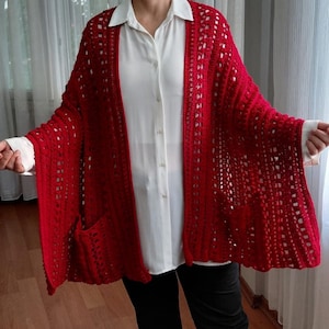 Puede incluir: Un cárdigan de ganchillo rojo brillante abierto por delante, con un patrón de puntos verticales texturizados y dos bolsillos delanteros. La prenda se lleva sobre una camisa blanca abotonada con cuello clásico y mangas largas.