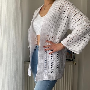 May include: A white, open-front crochet cardigan with long sleeves. The cardigan features a textured, open weave design. The garment is worn over a white top and blue jeans, suggesting a casual, layered outfit.