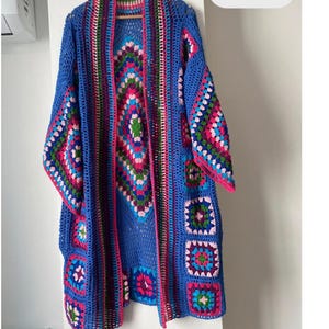 May include: A long, open-front crochet cardigan featuring a vibrant granny square pattern in shades of blue, pink, green, and purple. The garment is constructed with a large central diamond motif on the back and smaller square motifs along the sleeves.