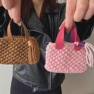 Könnte beinhalten: Zwei Miniaturhandtaschen aus strukturiertem Häkelstich. Eine Tasche ist braun mit beigen kunstlederartigen Griffen und einer passenden Quaste. Die andere Tasche ist hellrosa mit leuchtend rosa kunstlederartigen Griffen und einer rosa Quaste mit einer violetten Perle.