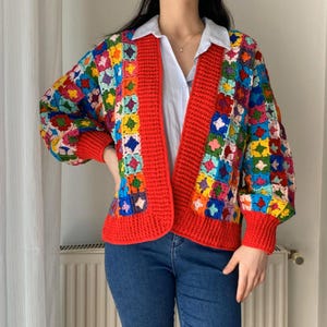 May include: A long-sleeve cardigan featuring a multi-colored granny square crochet pattern in shades of red, blue, yellow, green, and purple. The garment has a solid red ribbed trim along the front opening, cuffs, and hem, worn over a white shirt.