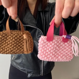 Op de afbeelding: Twee kleine gehaakte handtassen, een bruine en een roze, worden vastgehouden door een persoon. De bruine tas heeft een kwast.