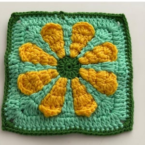 May include: A square, crocheted piece featuring a large yellow flower with green and yellow petals. The flower is centered on a light green background, with a dark green border. The crochet work is visible, showcasing the texture of the yarn.