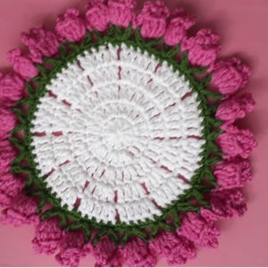May include: A handmade crochet doily featuring a central white circle with a radiating pattern. The doily is bordered by a green trim and pink crochet flower buds, creating a floral design. The background is a solid pink.