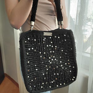 May include: A black, square-shaped handbag with a woven texture and silver sequins arranged in a radial pattern. The bag has thick, black rope handles with silver metal accents. A small silver label is centered on the top.