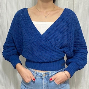 May include: A cobalt blue knitted wrap-over sweater with long sleeves and a V-neck. The sweater features a ribbed hem and cuffs. The model is wearing blue jeans and a white top. The sweater is a striking shade of blue.