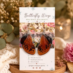 May include: Dangle earrings with butterfly wing designs. The wings are orange with dark brown spots, suspended from silver hooks. The product card reads "Butterfly Wings. Real Wings. Real Beauty."