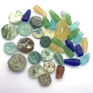 May include: A collection of assorted small glass beads in various shapes and colours, including shades of blue, green, yellow, and brown. The beads feature weathered, textured surfaces with some showing translucent qualities and central drill holes.