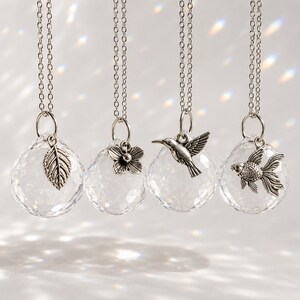 May include: Four clear crystal pendants with silver-colored charms. Charms include a leaf, flower, hummingbird, and goldfish. Each pendant hangs from a silver chain. The pendants are faceted, reflecting light.