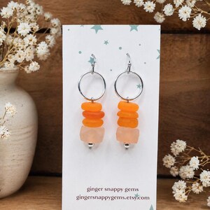 May include: A pair of dangle earrings with silver hoops. Each earring features a stack of orange and peach-colored sea glass beads. The earrings are displayed on a white card with the text "ginger snappy gems" and a website address.