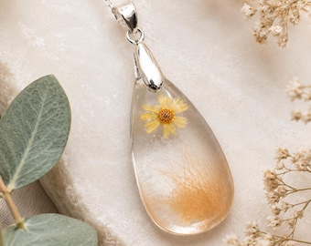 Könnte beinhalten: Ein tropfenförmiger Anhänger an einer Kette mit einem klaren Harzgehäuse, das eine kleine gelbe Blume und feine goldene Fasern umschließt. Der Anhänger ist an einer dünnen silberfarbenen Metallkette mit einer polierten silberfarbenen Öse befestigt.