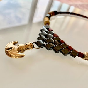 Könnte beinhalten: Eine goldfarbene Anhängerkette mit einem einzigartigen Design. Die Halskette hat einen goldfarbenen Ankeranhänger und eine geometrische Anordnung aus dunkelgrauen und roten Perlen. Die Halskette ist ein stilvolles Accessoire.