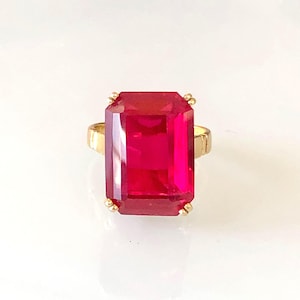 Peut inclure: Une bague dorée ornée d'une grande pierre précieuse rouge rubis de forme rectangulaire. La pierre précieuse est le point central, avec une surface à facettes qui capte la lumière. Le corps de la bague a un design simple et texturé.