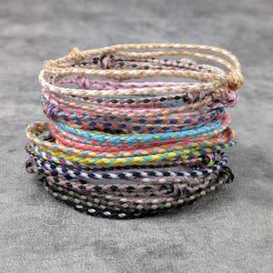 Könnte beinhalten: Ein Stapel geflochtener Freundschaftsarmbänder in verschiedenen Farben, darunter Beige, Rosa, Blau und Schwarz. Die Armbänder sind geknotet und scheinen aus einem gewebten Schnurmaterial zu bestehen. Die Armbänder sind übereinander gestapelt.