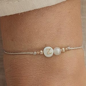 Könnte beinhalten: Ein zartes Armband mit einer neutralfarbenen Kordel zeigt einen weißen, kreisförmigen Anhänger mit dem Buchstaben "Z", einer Perle und goldfarbenen Perlen. Das Armband wird an einem Handgelenk gezeigt, mit dem Logo "ARTWAVES design".
