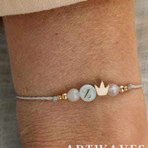 May include: A delicate bracelet with a light grey cord features a gold crown charm and a light blue bead with the letter "Z". Additional gold beads and a pearl-like bead are also present. The bracelet is a stylish accessory.