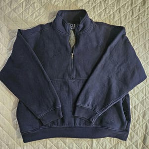 Puede incluir: Una sudadera azul marino con cremallera de un cuarto y la palabra "GAP" bordada en la parte delantera.