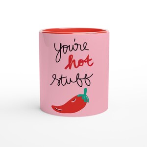 Könnte beinhalten: Rosa Keramiktasse mit rotem Rand. Die Tasse hat ein weißes Chili-Pfeffer-Motiv und den Text "you're hot stuff" in schwarzer Schrift.