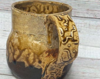 May include: A handmade ceramic mug with a brown and tan glaze. The mug has a unique, textured design and a large handle.