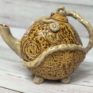 Può includere: Una teiera in ceramica marrone e beige con un design unico e testurizzato. La teiera ha un corpo arrotondato con un beccuccio e un manico curvi.