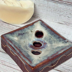 May include: A square ceramic soap dish with a white glaze and brown accents. The dish has three holes in the bottom for drainage.