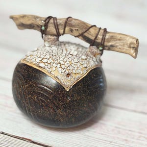 May include: A brown ceramic vase with a white crackle glaze and a natural wood handle. The handle is wrapped with brown leather cord and has a small green stone accent.