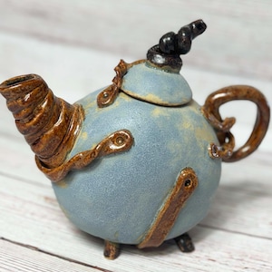 Peut inclure: Une théière en céramique bleue avec un bec et une poignée torsadés de couleur brune. La théière a un design unique et fantaisiste avec des accents bruns et un petit bouton noir sur le couvercle.
