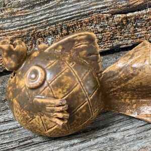 May include: A brown ceramic fish figurine with a textured surface and a large eye. The fish has a small fin on its head and a large tail fin.