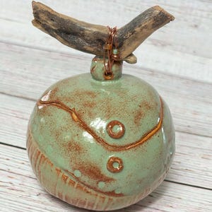 May include: A green ceramic jar with a rustic brown glaze and a driftwood lid. The jar has a textured surface with raised circles and lines. The lid is secured with a copper wire and beads.