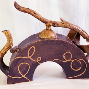 May include: A brown ceramic teapot with a unique design. The teapot is shaped like an arch with a branch for a handle. The teapot has a brown glaze with gold accents.