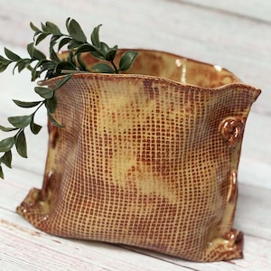 May include: A ceramic planter with a textured surface that resembles burlap. The planter is a light yellow color with brown accents. It has a wide opening and a rounded shape.