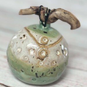 May include: A ceramic jar with a green, white and brown glaze. The jar has a round shape and a textured surface. A piece of driftwood is attached to the top of the jar.