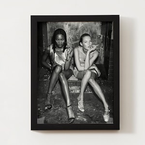 Puede incluir: Fotograf&iacute;a en blanco y negro enmarcada con dos mujeres sentadas, cada una sosteniendo un cigarrillo. Llevan vestidos y tacones. La fotograf&iacute;a est&aacute; en un marco negro, montada en una pared blanca.