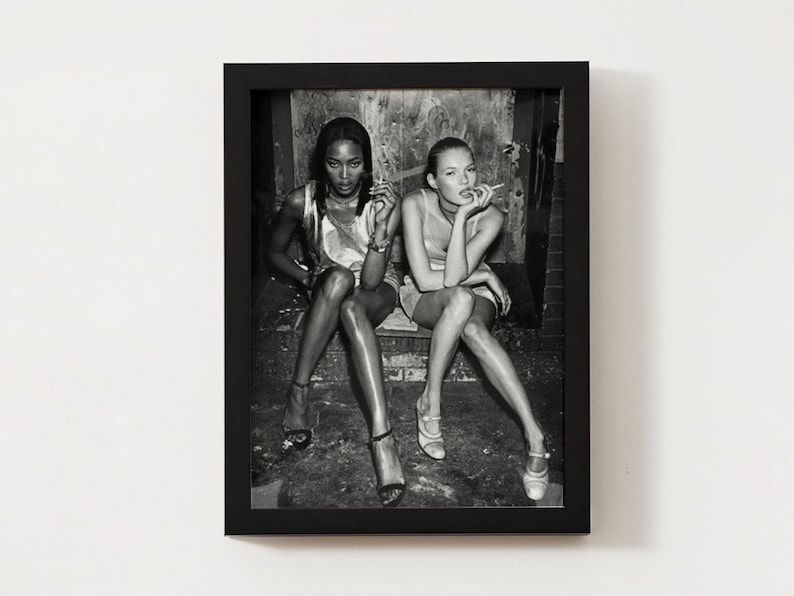 Puede incluir: Fotograf&iacute;a en blanco y negro enmarcada con dos mujeres sentadas, cada una sosteniendo un cigarrillo. Llevan vestidos y tacones. La fotograf&iacute;a est&aacute; en un marco negro, montada en una pared blanca.