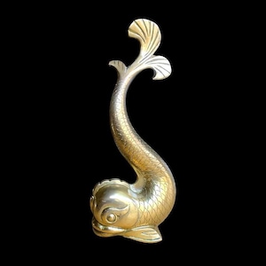 Puede incluir: Una escultura de pez estilizada de color latón, con un cuerpo curvado y una aleta caudal trilobulada. El pez tiene escamas detalladas y una cabeza redondeada. La escultura está sobre un fondo negro.