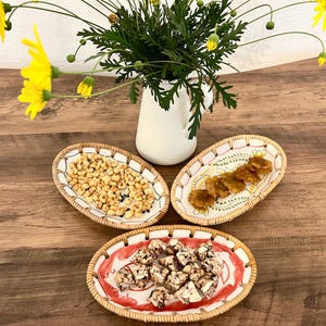 May include: Three oval-shaped ceramic serving dishes with decorative designs. The dishes are filled with snacks, including small, light brown, puffed corn kernels, small, light brown, chewy candies, and small, dark brown, chewy candies.