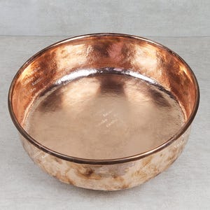 May include: A large, round copper bowl with a hammered finish. The bowl is empty and has a shiny, reflective surface.
