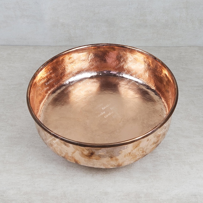 May include: A large, round copper bowl with a hammered finish. The bowl is empty and has a shiny, reflective surface.