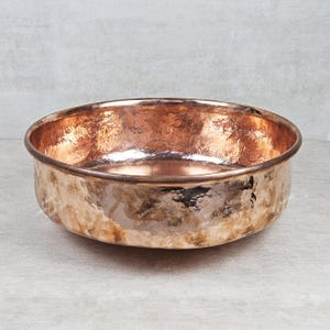 May include: A large, round copper bowl with a hammered finish. The bowl is empty and has a shiny, metallic finish.