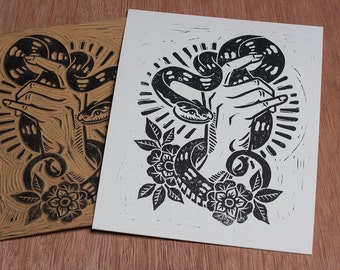 May include: Black and white linocut print of a hand holding a snake with a floral design. The snake is coiled around the hand and its head is facing the viewer. The print is on a white background.
