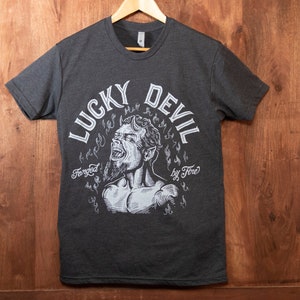 May include: A dark heather grey t-shirt with a white graphic print of a devil with horns and flames. The text "Lucky Devil" and "Forged by Fire" are printed in a white circular design around the devil.