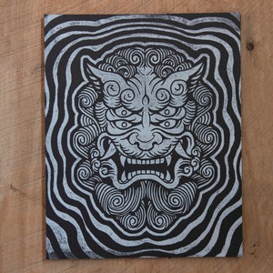 May include: A black and white illustration of a Japanese Oni mask with swirling patterns. The mask has a fierce expression with large eyes, a wide mouth, and sharp teeth.